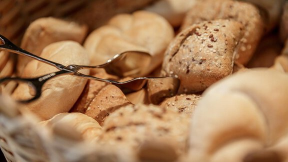 A basket with various rolls, including light and dark bread. A pair of tongs is ready to serve the rolls. - Apart Garni Enzian - Sölden | © RudiWyhlidal