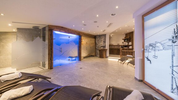 A modern wellness area with loungers and soft lighting. In the background, there is a glass wall with blue light and a view of the mountains. - Apart Hotel Garni Alt Kaisers - Sölden