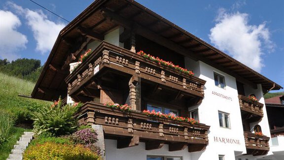 A charming alpine building with blooming balconies and a well-maintained garden. The clear sky and mountains in the background create an inviting atmosphere. - Appartement Harmonie - Sölden