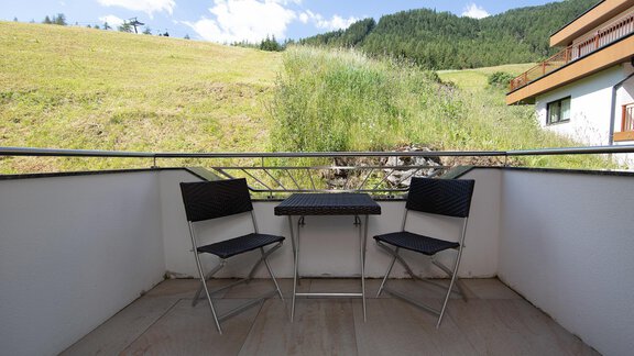 A balcony with two black chairs and a small table. In the background, a green meadow and wooded hills stretch under a blue sky. - Appartement Harmonie - Sölden