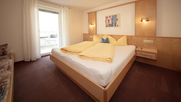 A modern bedroom with a large bed and yellow pillows. The windows let in a lot of light and provide access to a balcony. - Appartement Harmonie - Sölden