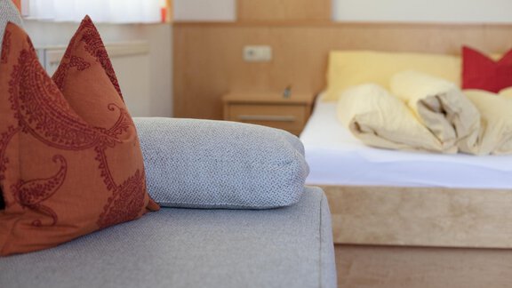 In the picture, there is a cozy corner with a gray sofa and colorful cushions. In the background, there is a bed with messy beddings. - Appartement Harmonie - Sölden