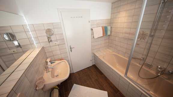 A modern bathroom with a bathtub and a shower. The walls are tiled with light tiles and there is a sink with a mirror. - Appartement Harmonie - Sölden