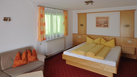 A cozy hotel room with a large bed and yellow pillows. A sofa with orange pillows and a table complement the decor. - Appartement Harmonie - Sölden