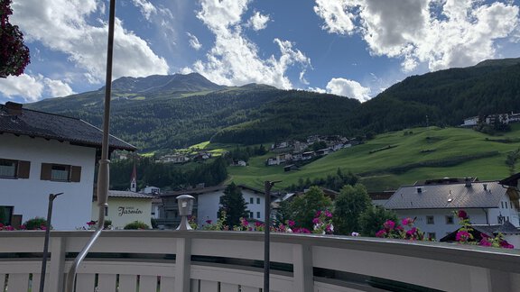 A picturesque mountain landscape with green hills and clouds in the blue sky. In the foreground, colorful flowers are visible in front of a balcony. - Appt. Ideal - Sölden