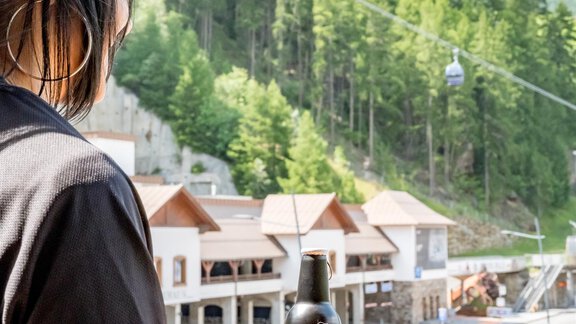 Eine Person steht auf einem Balkon und betrachtet die Landschaft mit Bergen und Bäumen. In der Hand hält sie eine Flasche Getränk. - Bäckelar Wirt - Sölden