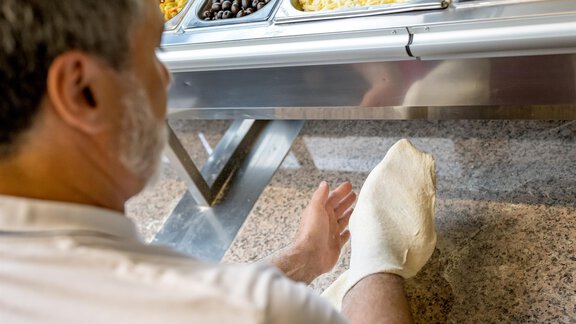 Ein Mann sitzt an einem Buffet und zeigt mit seinem Fuß auf einen Tisch. Auf dem Buffet sind verschiedene Lebensmittel in Schalen angeordnet. - Bäckelar Wirt - Sölden