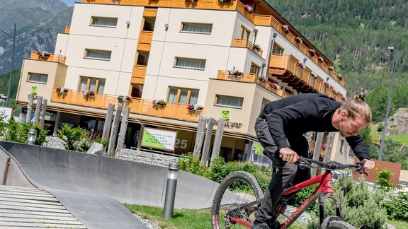 Ein Radfahrer springt über eine Rampe in einer grünen Umgebung. Im Hintergrund sieht man ein Gebäude und Berge unter einem blauen Himmel. - Bäckelar Wirt - Sölden