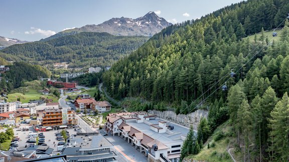 Eine malerische Berglandschaft mit grünen Wäldern und schneebedeckten Gipfeln. Im Vordergrund sind moderne Gebäude und eine Seilbahn zu sehen. - Bäckelar Wirt - Sölden