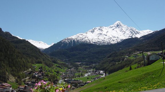 A beautiful mountain landscape with snowy peaks and a clear blue sky. In the foreground, green meadows and a small village are visible. - Brandlehof - Sölden