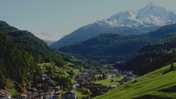 A picturesque mountain landscape with snow-capped peaks and green meadows. In the foreground, you can see a small village surrounded by gentle hills. - Brandlehof - Sölden
