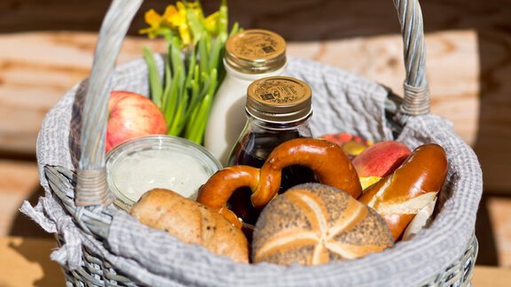 A woven basket with various foods: apples, pretzels, milk, and honey. Fresh spring flowers are visible in the background. - Chalet Resort Sölden - Sölden