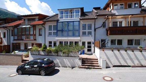 A modern building on a quiet street with several floors. In front of the house, there is a car, and the surroundings are characterized by greenery and beautiful houses. - Ferienhaus Moidl - Sölden