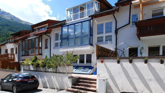 A modern residential building with large windows and a friendly entrance. In the foreground, there is a parked car and there are some plants in the garden. - Ferienhaus Moidl - Sölden