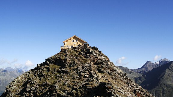A small, yellow house stands on a rocky peak. In the background, majestic mountains can be seen under a clear, blue sky. - Gästehaus Europa - Sölden