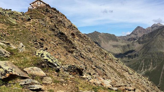 A steep mountainous landscape with grassy vegetation and rocks. In the background, a small house is visible on a peak, surrounded by mountains and blue sky. - Gästehaus Martin - Sölden