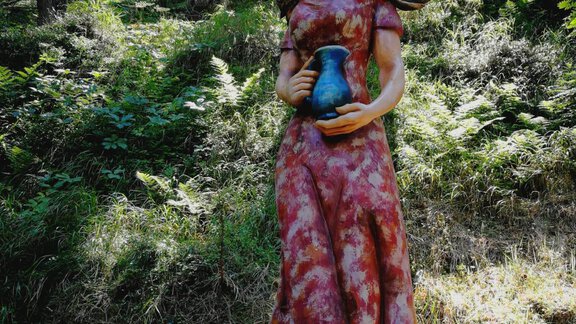 A colorful statue of a woman in a long dress holds a jug. She stands in a wooded setting with green forest in the background. - Gästehaus Martin - Sölden
