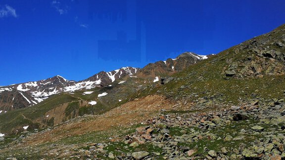 A breathtaking mountain landscape with snow-capped peaks and a clear blue sky. In the foreground, green meadows and rocks are visible. - Gästehaus Martin - Sölden