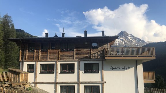 A modern building with wooden cladding, surrounded by mountain scenery. In the background, snow-covered mountains and a blue sky can be seen. - Granbichlhof - Sölden