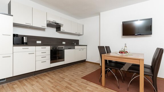 A modern kitchen with white cabinets and a cooking area. In the foreground, there is a table with chairs and a television on the wall. - Gurgler Hof - Sölden