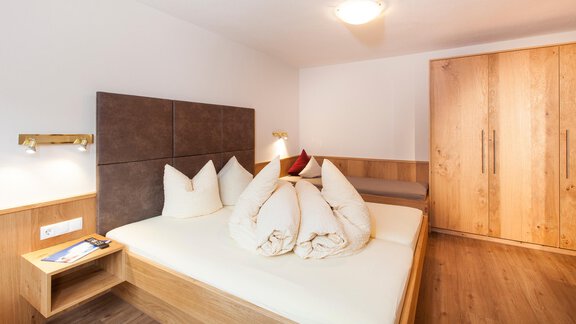 A cozy bedroom with a double bed and decorative cushions. The room features a wooden wall and a wardrobe made of light wood. - Haus Bergblick - Sölden