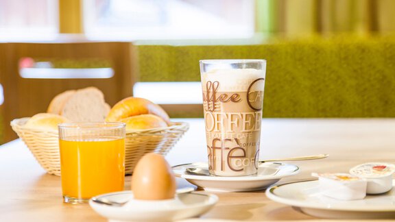 A laid breakfast table with a glass of coffee, a boiled egg, and a glass of orange juice. In the background, there is a basket with rolls. - Haus Bergblick - Sölden