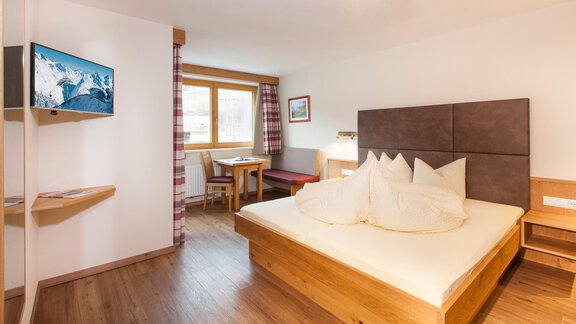 A cozy bedroom with a large bed and a television on the wall. There is a table and a chair, as well as a window with a view outside. - Haus Bergblick - Sölden
