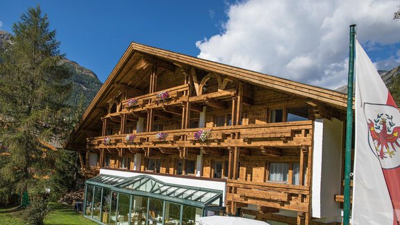 A charming wooden building in the mountains with blooming balcony flowers. In the foreground, a sunny conservatory is visible. - Hotel am Hof - Sölden