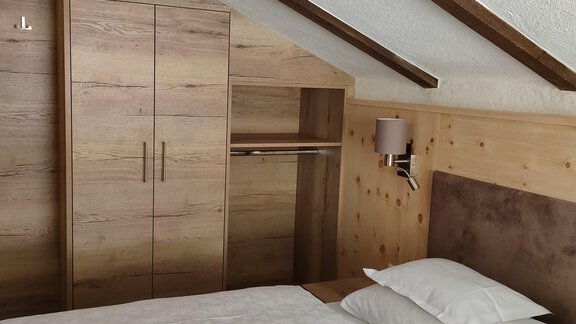 A cozy room with a bed and wooden paneling. There is a built-in wardrobe and a bedside lamp. - Hotel am Hof - Sölden