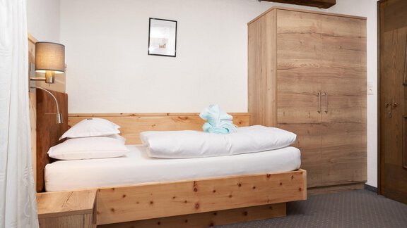 A simple bedroom with a wooden bed and white bedding. A small picture hangs on the wall, and there is a wooden wardrobe in the room. - Hotel am Hof - Sölden