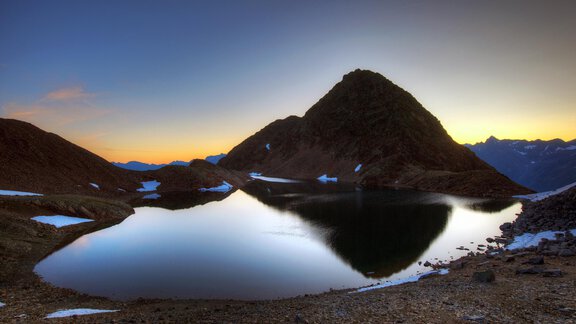 Eine ruhige Berglandschaft mit einem klaren See und schneebedeckten Flächen. Im Hintergrund ist ein majestätischer Gipfel bei Sonnenuntergang zu sehen. - La Luna - Sölden
