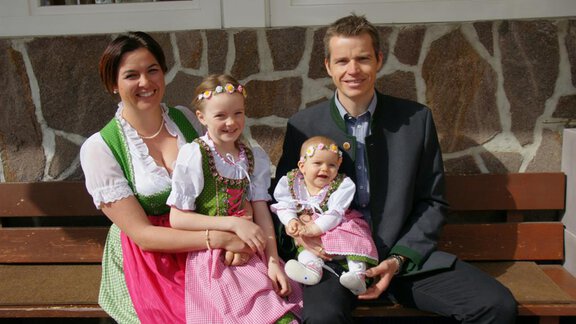 A family is sitting on a bench and wearing traditional Bavarian costumes. There are two girls and one adult, all smiling kindly at the camera. - Pension Sportalm - Sölden