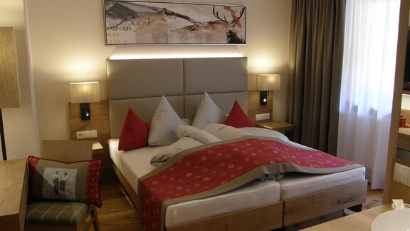 A cozy bedroom with a double bed and red pillows. The room is bright and modernly furnished, with a large window and wooden details. - Pension Sportalm - Sölden