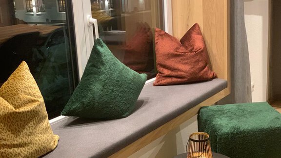 A cozy window seat with various cushions in warm colors. Next to the cushions is a small table with a decorative lantern. - Pension Sportalm - Sölden