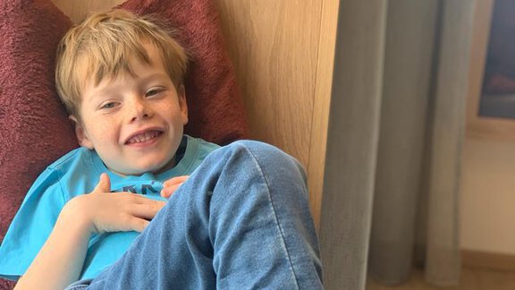 A cheerful boy is sitting relaxed on a sofa and smiling. He is wearing a light blue t-shirt and comfortable jeans. - Pension Sportalm - Sölden