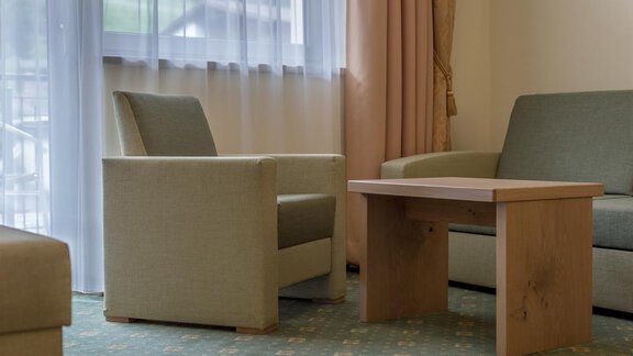 A modern living room with a light carpet and neutral furniture. There is a table and two comfortable seating options next to a window with curtains. - Residenz Allegra - Sölden | © Residenz Allegra Sölden