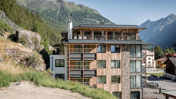 A modern building with wooden elements and large windows in a mountainous landscape. In the background, majestic mountains can be seen. - Valentin Design Apartment - Sölden