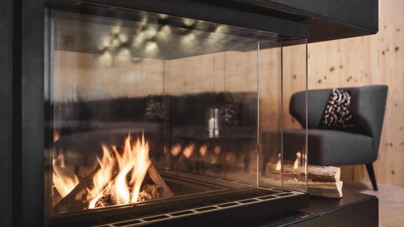 A modern fireplace with a clear glass front that displays a warm fire. In the background, a cozy armchair can be seen. - Valentin Design Apartment - Sölden