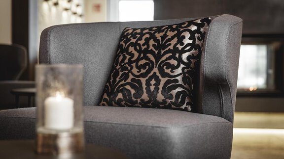 An elegant gray armchair with a decorative cushion. In the background, a cozy fireplace is visible. - Valentin Design Apartment - Sölden