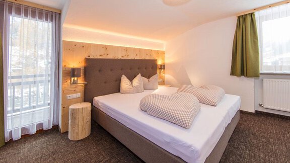 A modern bedroom with a large bed, decorative pillows, and soft lighting. The windows let in plenty of daylight and offer a cozy view outside. - Vizensn Living - Sölden