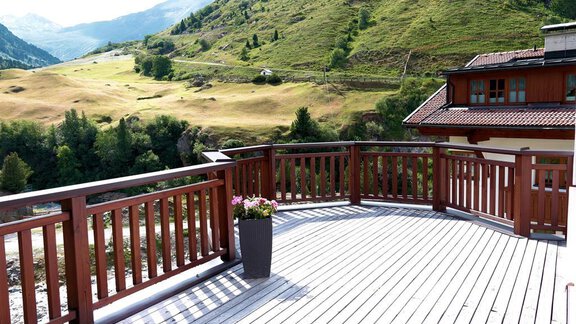 A terrace with a wooden railing overlooking green hills. In the foreground is a flower pot with flowers. - Gästehaus Soldanella - Vent