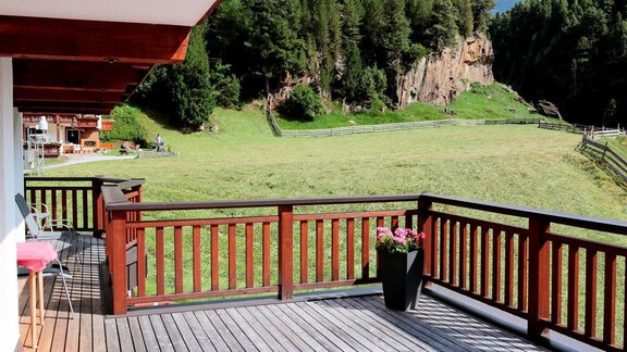 A terrace with a wooden railing and a view of a green meadow. In the background, trees and rocks can be seen under a clear blue sky. - Gästehaus Soldanella - Vent