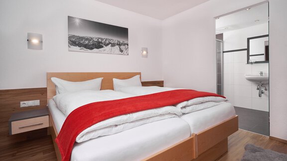 A modern bedroom with a large bed and a red blanket. The room is bright and friendly with a view of a bathroom. - Apart Alessandro - Huben