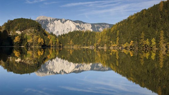Eine ruhige Landschaft mit einem klaren See, umgeben von Bäumen und Bergen. Die Spiegelung der Berge ist deutlich im Wasser zu sehen. - Appartement Erich Wilhelm - Huben