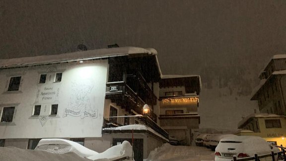 A snowy landscape at night with two buildings and some cars. The snow is deep and it appears to be snowing just now. - Haus Verwall Appartements - Obergurgl 