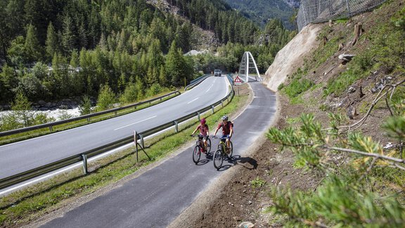 Zwei Fahrradfahrer fahren auf einem Radweg neben einer Straße. Die Umgebung ist grün und bergig mit einer Brücke im Hintergrund. - Gasthof Waldesruh - Ochsengarten