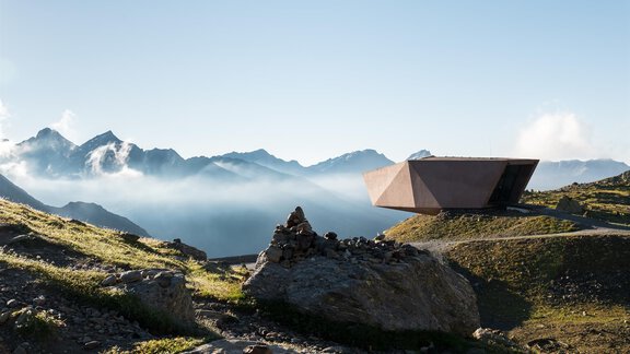 Ein modernes, geometrisches Gebäude steht in einer beeindruckenden Berglandschaft. Der Himmel ist klar und die Berge sind mit einer leichten Nebelschicht umhüllt. - Gasthof Waldesruh - Ochsengarten