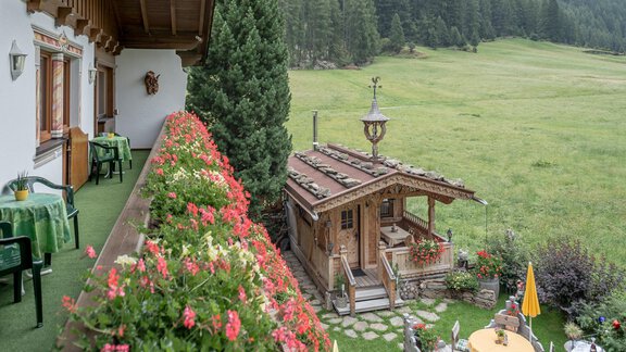 A cozy balcony with colorful flowers and a view of a green meadow. In the background stands a rustic wooden house, surrounded by trees and mountain scenery. - Apart Wiesengrund - Sölden