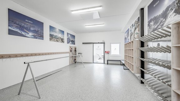 A bright room with a gray floor and white walls. Pictures of mountains hang on the wall, and there are shelves as well as a pole in the middle of the room. - Appartementhaus Kathrin - Sölden