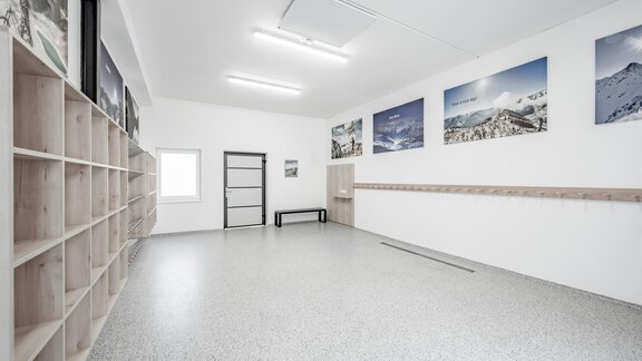 A bright room with white walls and gray flooring. Many photos of mountains hang on the wall, and there are shelves as well as a bench. - Appartementhaus Kathrin - Sölden